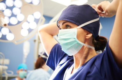 Side view of a female surgeon in navy blue scrubs and a cap ties her mask as she prepares for surgery.