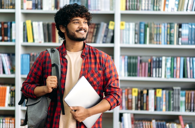 A man in a red checkered shirt stands with a backpack over one shoulder and holds a laptop in a library.