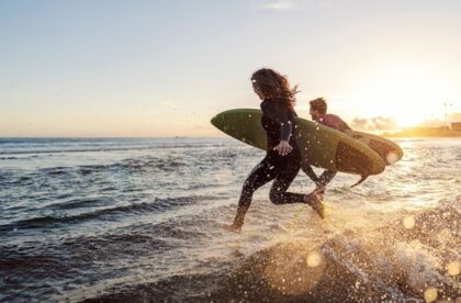 A man and a woman run into the ocean with their surfboards as the sun goes down. They're both wearing wetsuits.