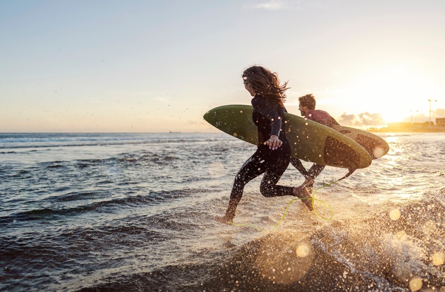 A man and a woman run into the ocean with their surfboards as the sun goes down. They're both wearing wetsuits.