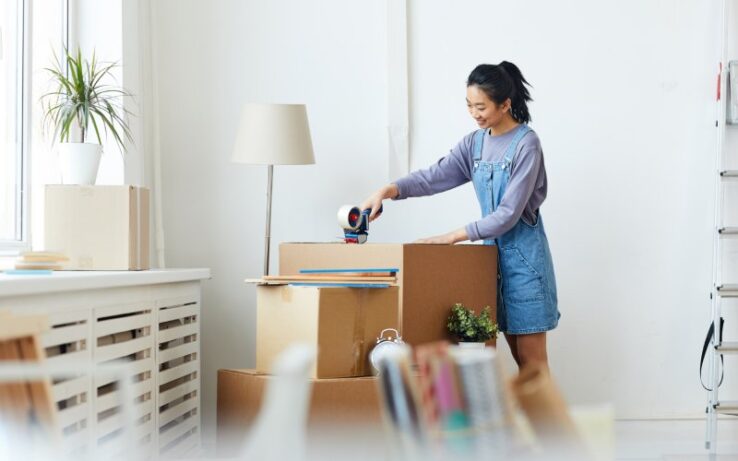 A young person using a tape roller to apply packaging tape to a large cardboard box while standing in a dorm room.