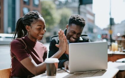Two young people sitting at an outdoor table and high fiving each other while both looking at a laptop screen.