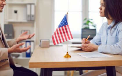 A man and a woman sit across from each other at a wooden table with an American flag. The woman is writing on paperwork.