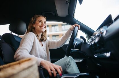 A young woman sits in the driver's seat of a car with one hand on the wheel as she smiles out the window.