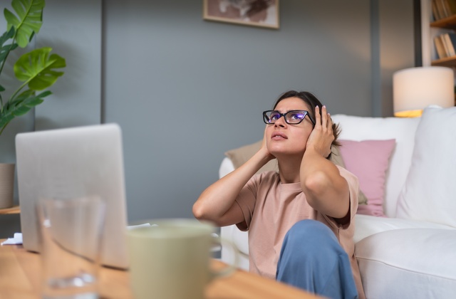 A college student wearing glasses sitting in front of their couch while trying to work, but excessive noise is distracting them.