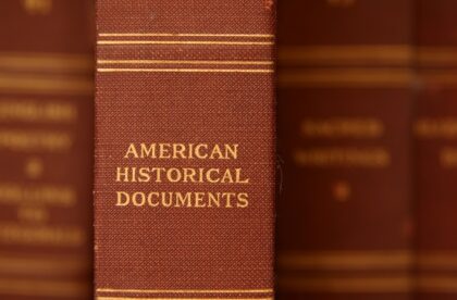 A close-up view of an old red book titled American Historical Documents on a bookshelf. There are other red books out of focus.