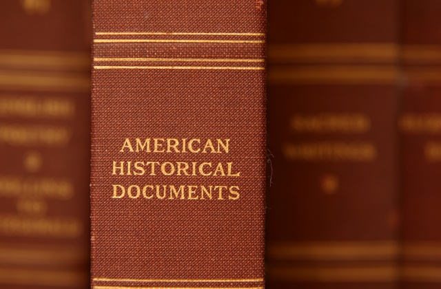 A close-up view of an old red book titled American Historical Documents on a bookshelf. There are other red books out of focus.
