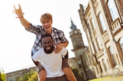 A Black college student carrying his White fraternity brother on his back as they smile big for the camera.