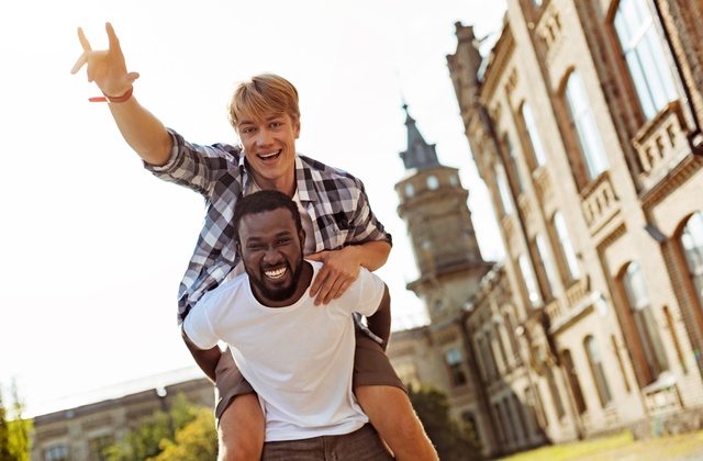 A Black college student carrying his White fraternity brother on his back as they smile big for the camera.