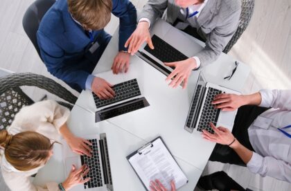 A group of students in professional business attire gathered around a table. They are working on their laptops.