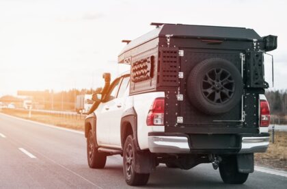A white pickup truck with a black cargo box and several accessories attached to the back as it drives down the road.