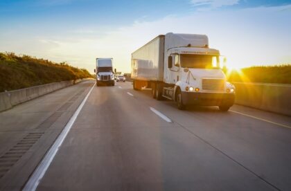 Two large, white semi trucks driving along a highway at sunset with passenger cars driving behind them.