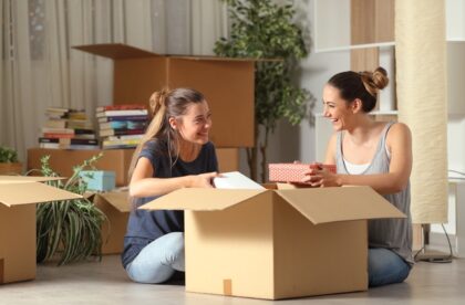 Two roommates laugh as they work together to unpack a box. They sit surrounded by boxes, books, and plants.