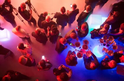 An overview of a cocktail party. People are standing at black tables underneath the pink and purple neon lights.