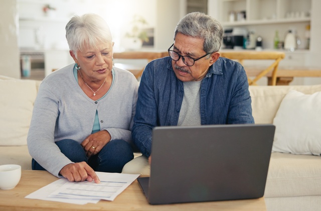 An older woman pointing to a document while sitting beside a man using a laptop on a coffee table in a bright living room.