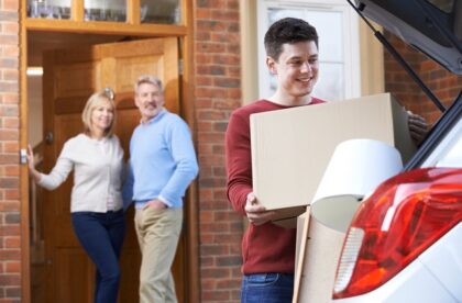 A close-up showing a young man in a red shirt loading a box into the trunk of a car as two adults watch him.