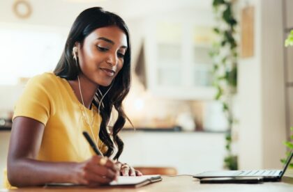 A young woman, wearing wired headphones, is using her laptop to write notes in her notebook at home.