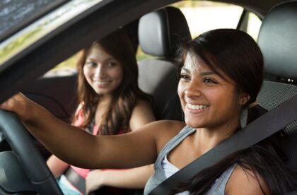 Two young women are driving in a car for a road trip and smiling. The interior of the vehicle is black.