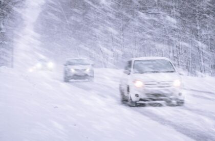 Multiple vehicles driving through a blizzard that is obscuring the road. Around the road are snow-covered trees.