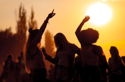 Silhouettes of several people together dancing outside against the sky that is orange in color due to the sunset.