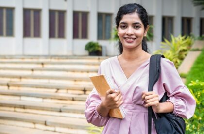 A young woman wearing a pale pink blouse smiles while holding a notebook and a backpack on a college campus.