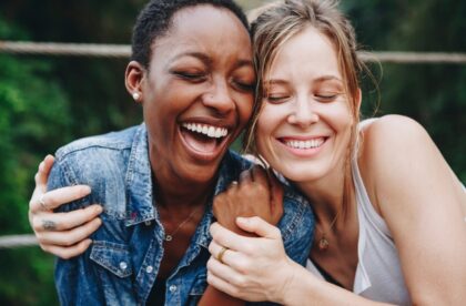 Two smiling women hug outdoors with their eyes closed. One wears a white tank top, and the other is in denim.