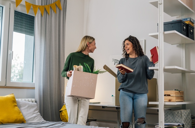Two college students put their books, decor, and other personal items on a shelving unit in their dorm room.
