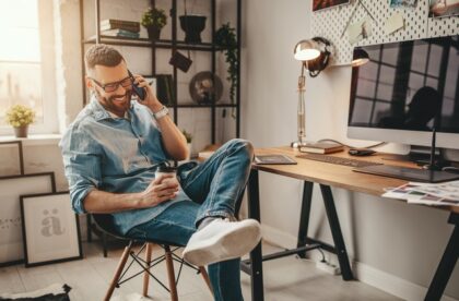 A young man talking on the phone while holding coffee. He's sitting in a modern home office with a computer and desk near him.
