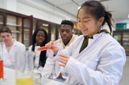 A group of students in white lab coats watching as one student pours a red liquid into a tube with a blue liquid.