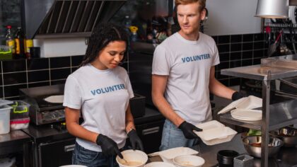 A young woman and a young man wearing white short-sleeved t-shirts that have the word "volunteer" written on them are preparing takeaway meals in a kitchen.