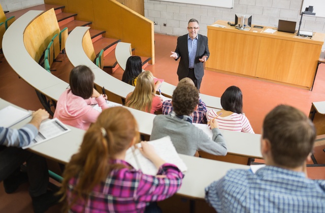 A middle-aged man, dressed in a suit and wearing glasses, faces a group of young adults with notebooks in a lecture hall.