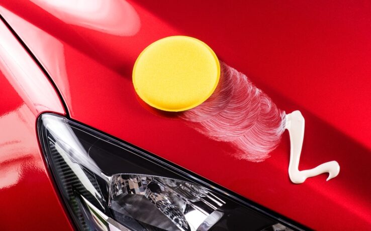 A car with red paint has a sponge on it, there is a streak of foam trailing behind the sponge on the car.