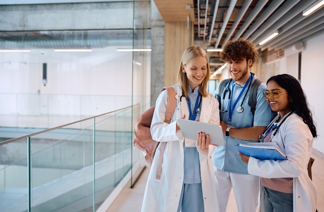 A group of medical students stand in a breezeway wearing backpacks and stethoscopes. They're looking at a tablet and smiling.