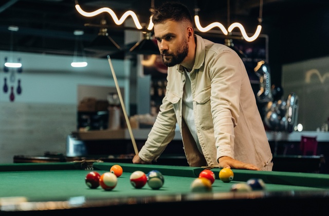 A guy thinking as he looks over a pool table with balls spread across it. He's leaning with both hands on the edge of the table.