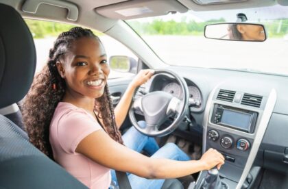 A teenage girl sits in the driver's seat of a vehicle. She is turning around and smiling with her hand on the gear shift.