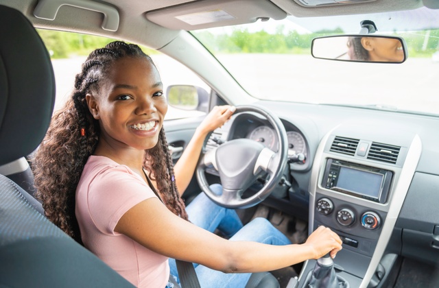 A teenage girl sits in the driver's seat of a vehicle. She is turning around and smiling with her hand on the gear shift.