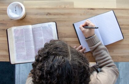 Someone sits at a desk with a religious text and a journal open in front of them. They write something down in the journal.