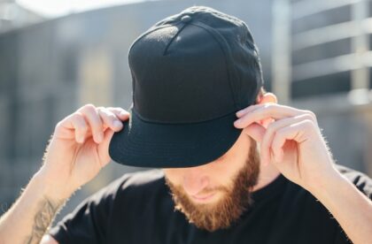 A close-up of the head of a man with a long brown beard, wearing a black baseball cap while holding it with both hands.