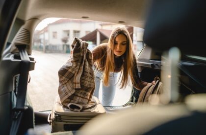 A woman puts her two suitcases and coat into the back of a van. The back hatch of the vehicle is open.