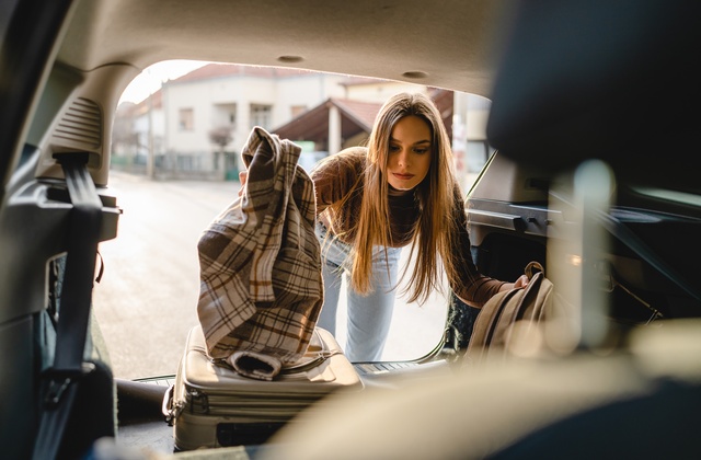 A woman puts her two suitcases and coat into the back of a van. The back hatch of the vehicle is open.