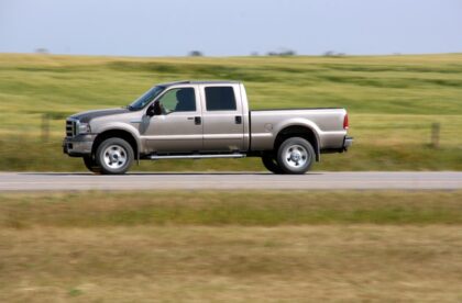 A pickup truck driving down the road during the daytime. The truck is driving past a large green field.