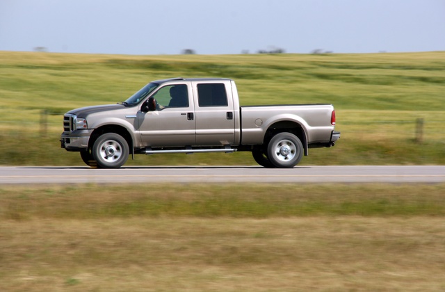 A pickup truck driving down the road during the daytime. The truck is driving past a large green field.