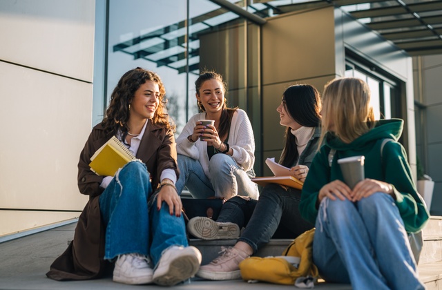 A group of college students in cold-weather attire sitting down on a step together and enjoying to-go drinks with each other.
