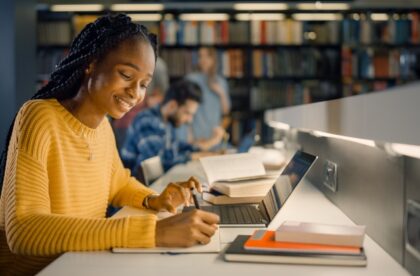 A college student sitting at a library desk area and working between a paper journal and a laptop, with books around them.