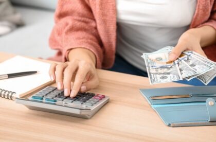 A close-up of a young woman, wearing a coral cardigan, is sitting at the table, counting money at home with a calculator.