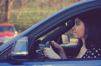 A woman in a black, polka-dotted shirt sits in the driver's seat of a car with a frustrated look on her face.