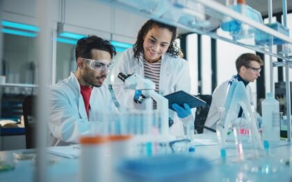 Three scientists in a lab collaborate around a microscope; one holds a tablet, and another works independently.