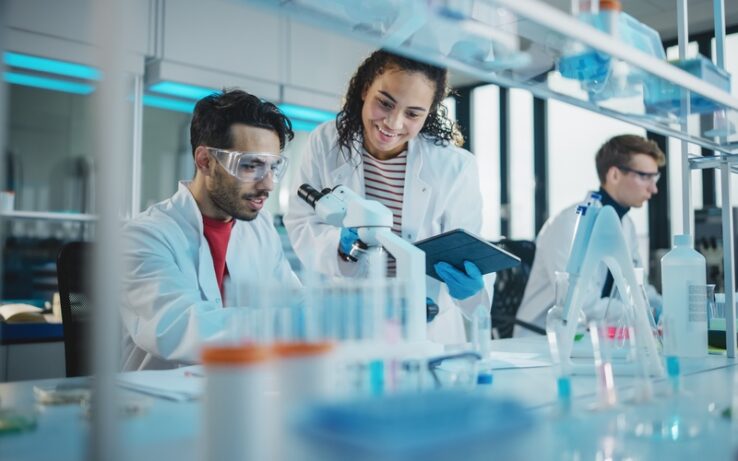 Three scientists in a lab collaborate around a microscope; one holds a tablet, and another works independently.