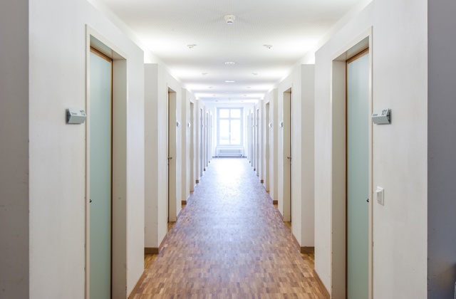 A long, bright dormitory hallway with doors on either side. Each door has a placard outside indicating the room number.