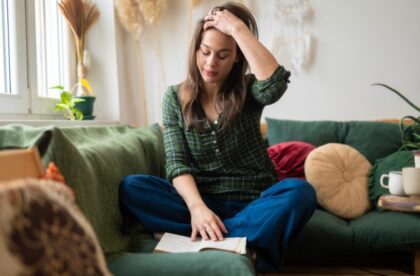 A young college student sits cross-legged on a green couch, holding her head while reading a book in her living room.